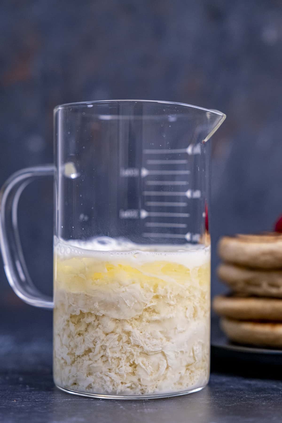 Dairy-free buttermilk in a glass measuring cup and pancakes behind it on a dark background.