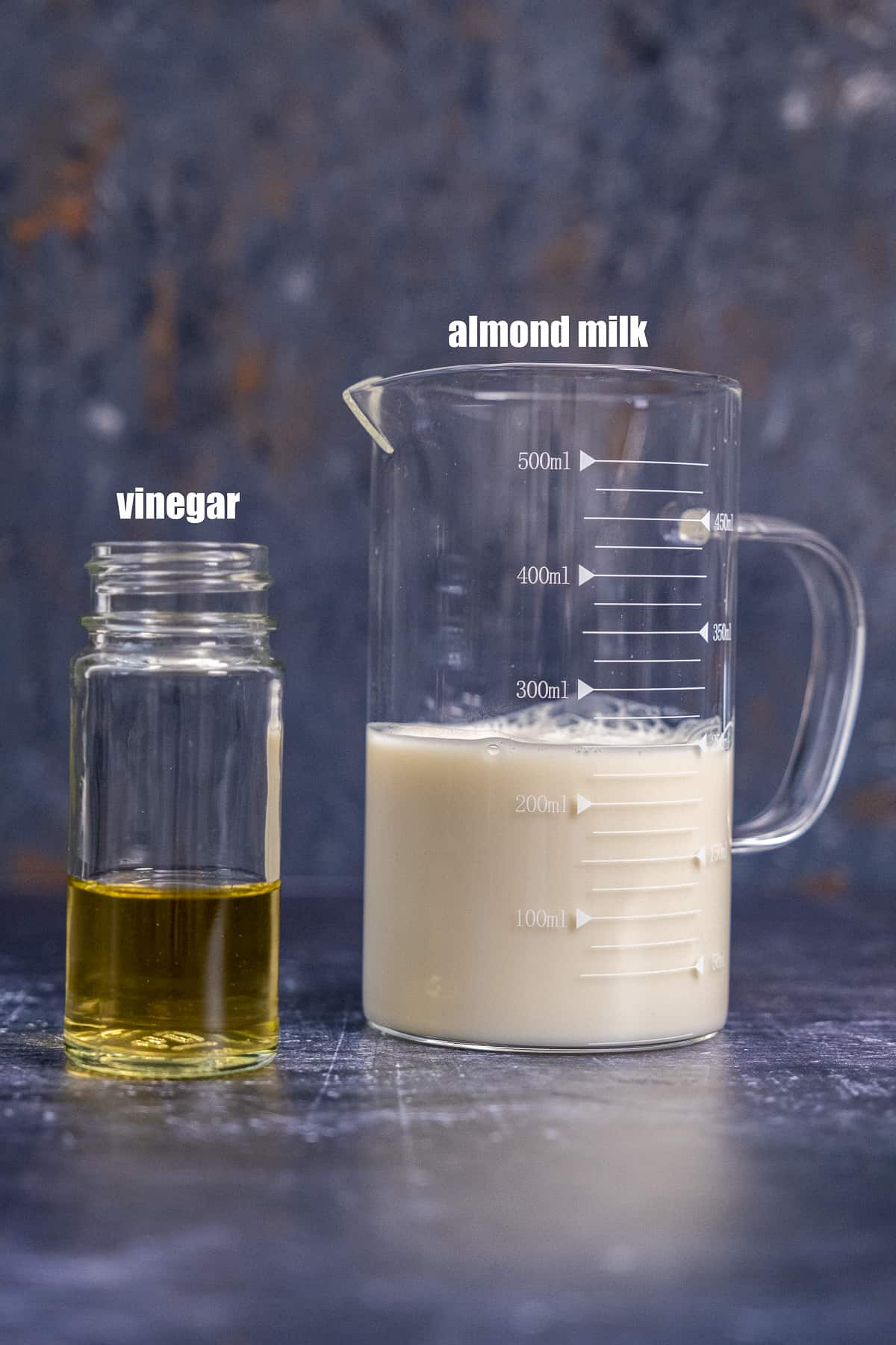 Almond milk in a glass measuring cup and apple cider vinegar in a small jar on a dark background.