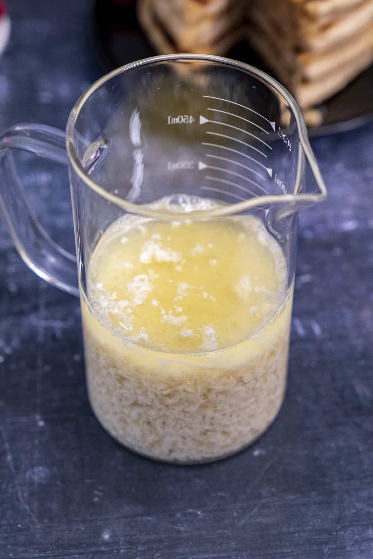 Dairy-free buttermilk in a glass cup and pancakes behind it on a dark background.