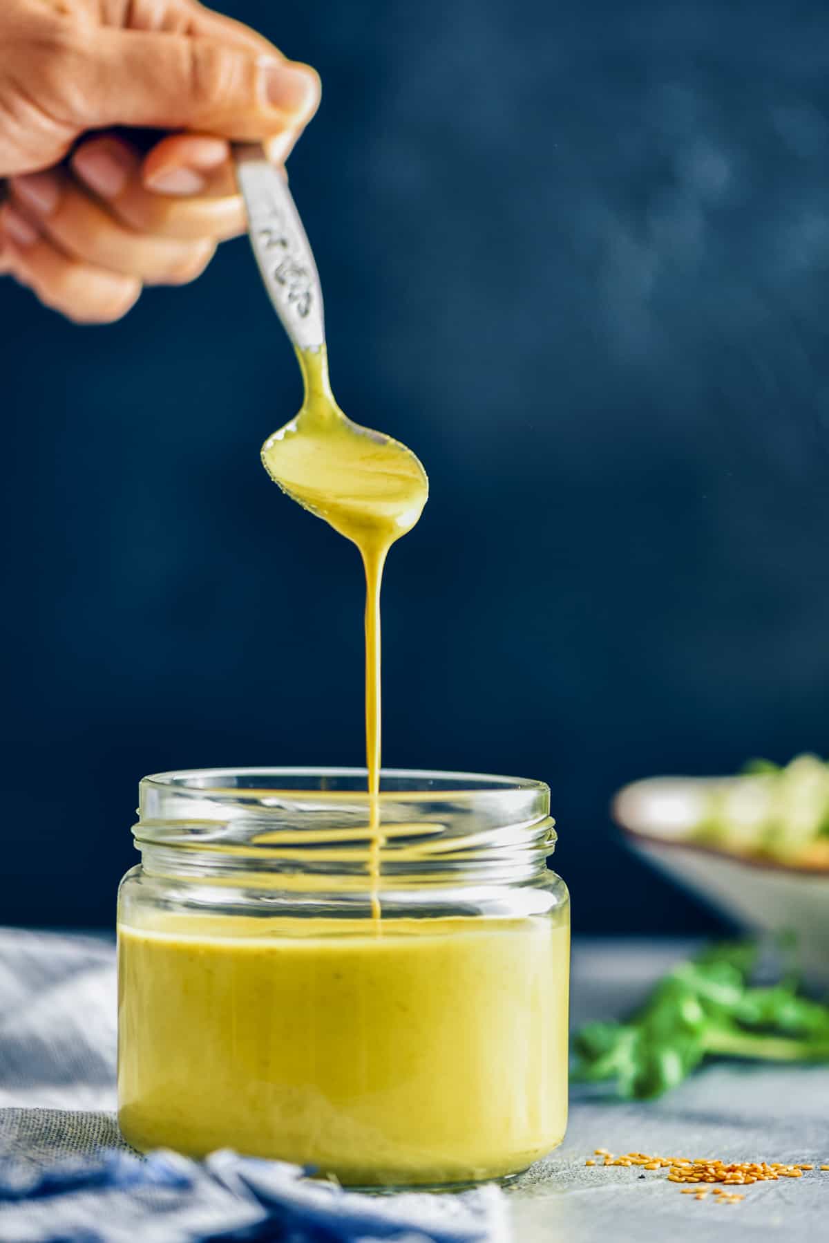 A hand holding a spoon dripping tahini dressing into a jar.