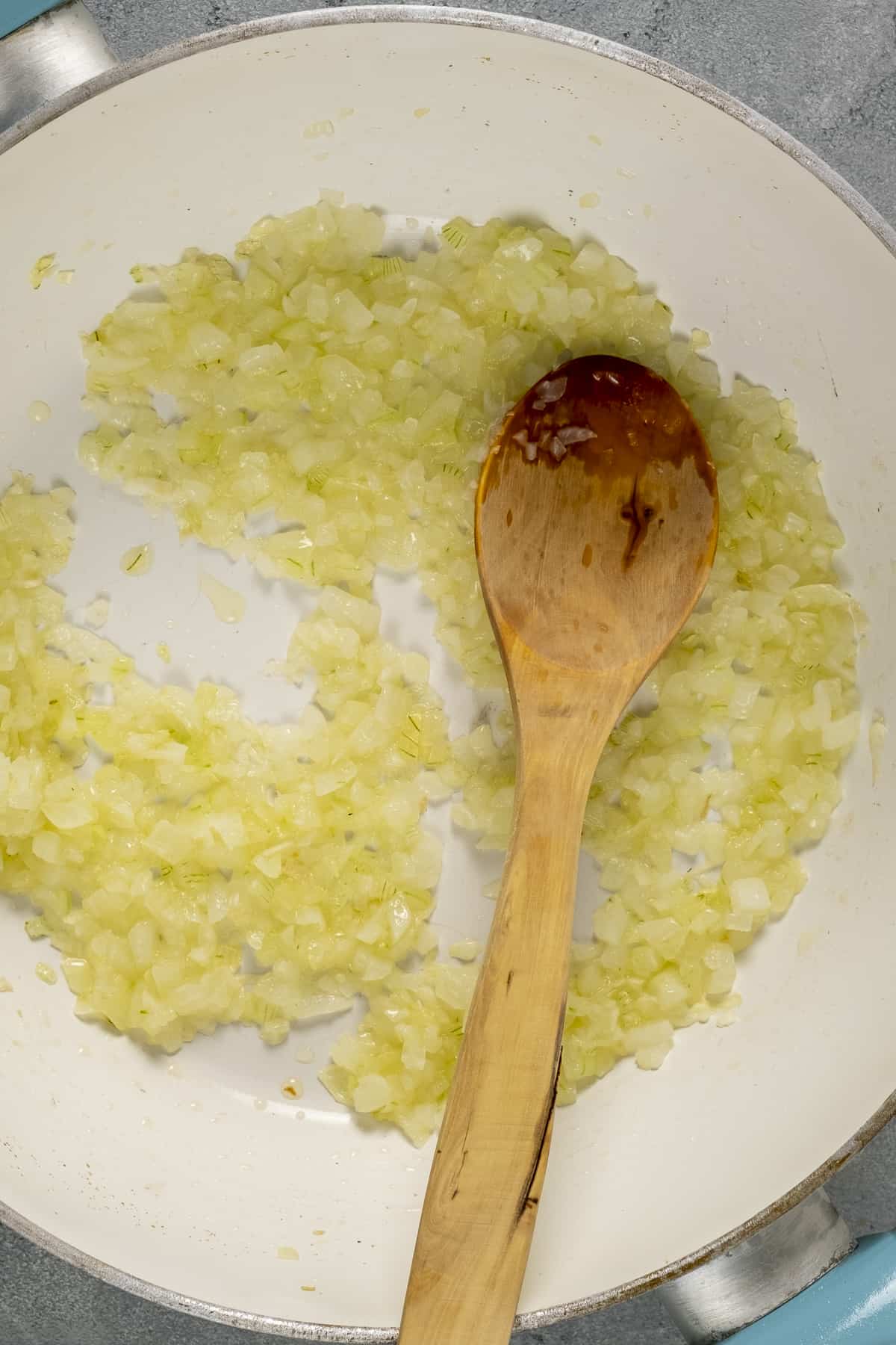 Finely chopped onions cooking in a white pan and a wooden spoon inside.
