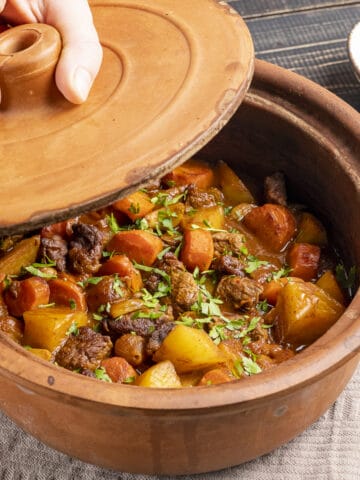 A hand opening the lid of a clay pot filled with stew.