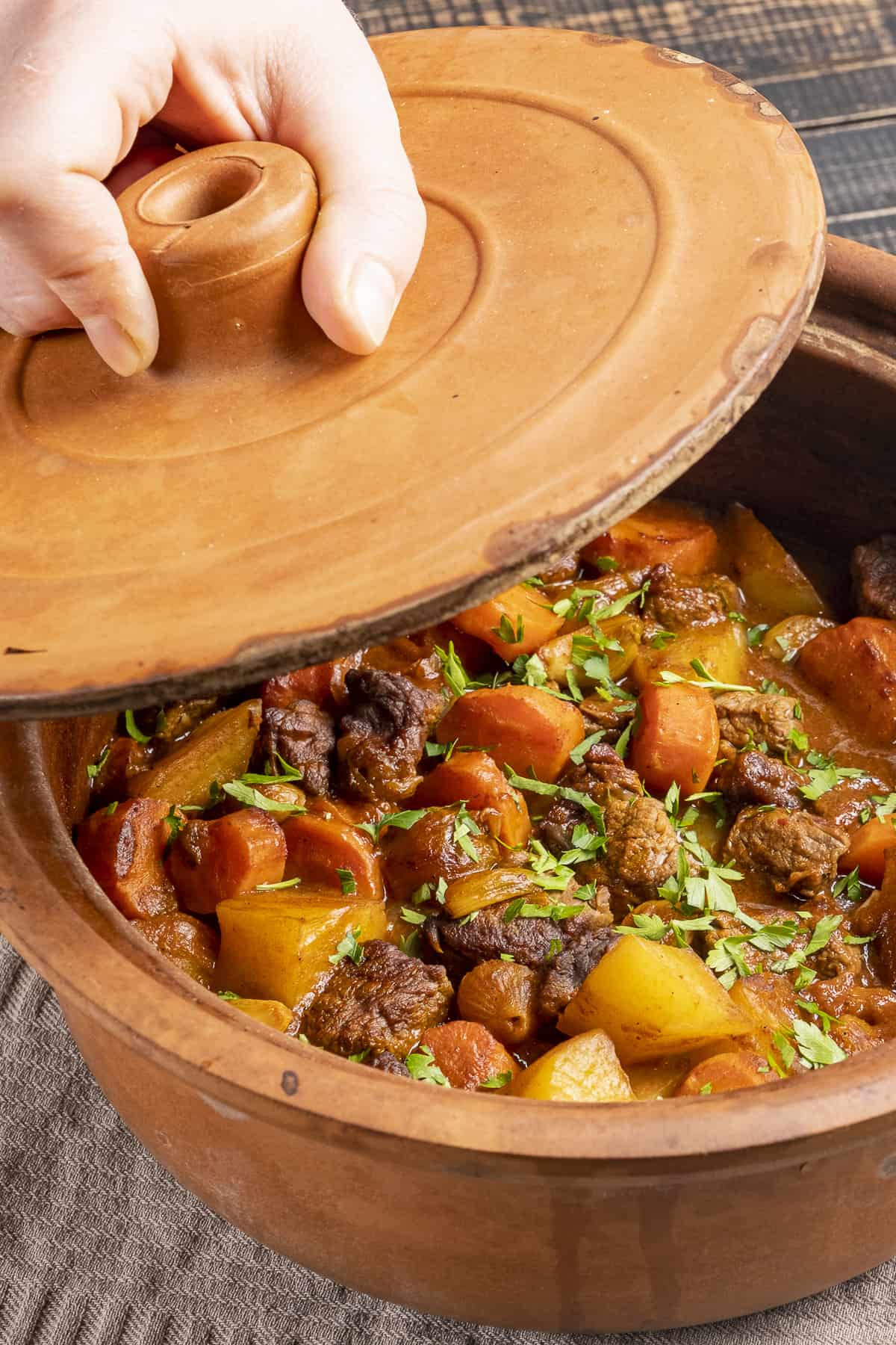 A hand opening the lid of a clay pot which is full of a stew.