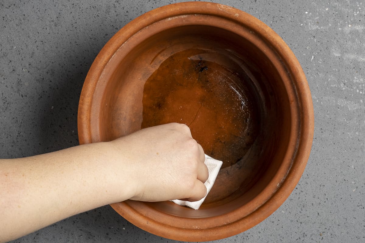 A hand oiling a clay pot using a piece of paper towel and oil.