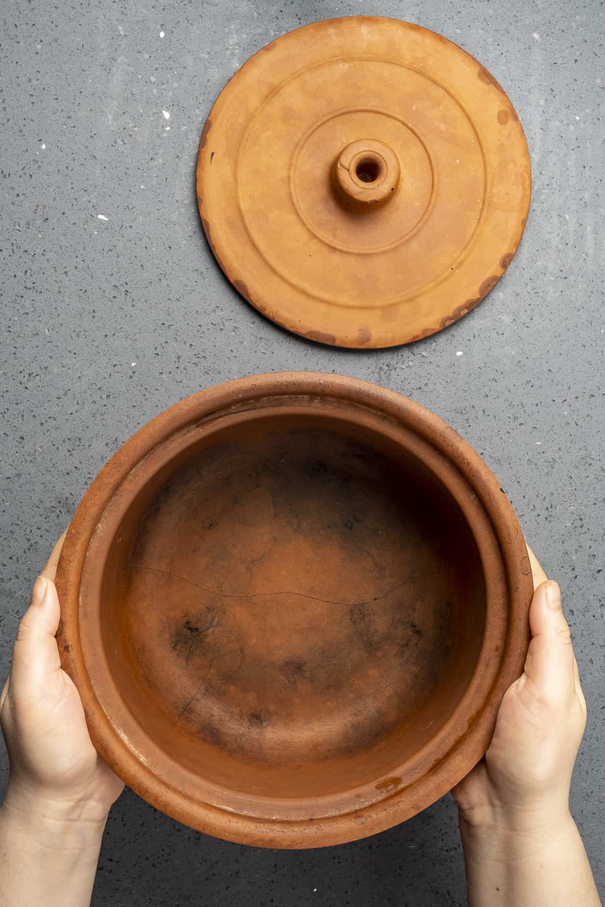 Hands holding a clay pot and a lid on the side.