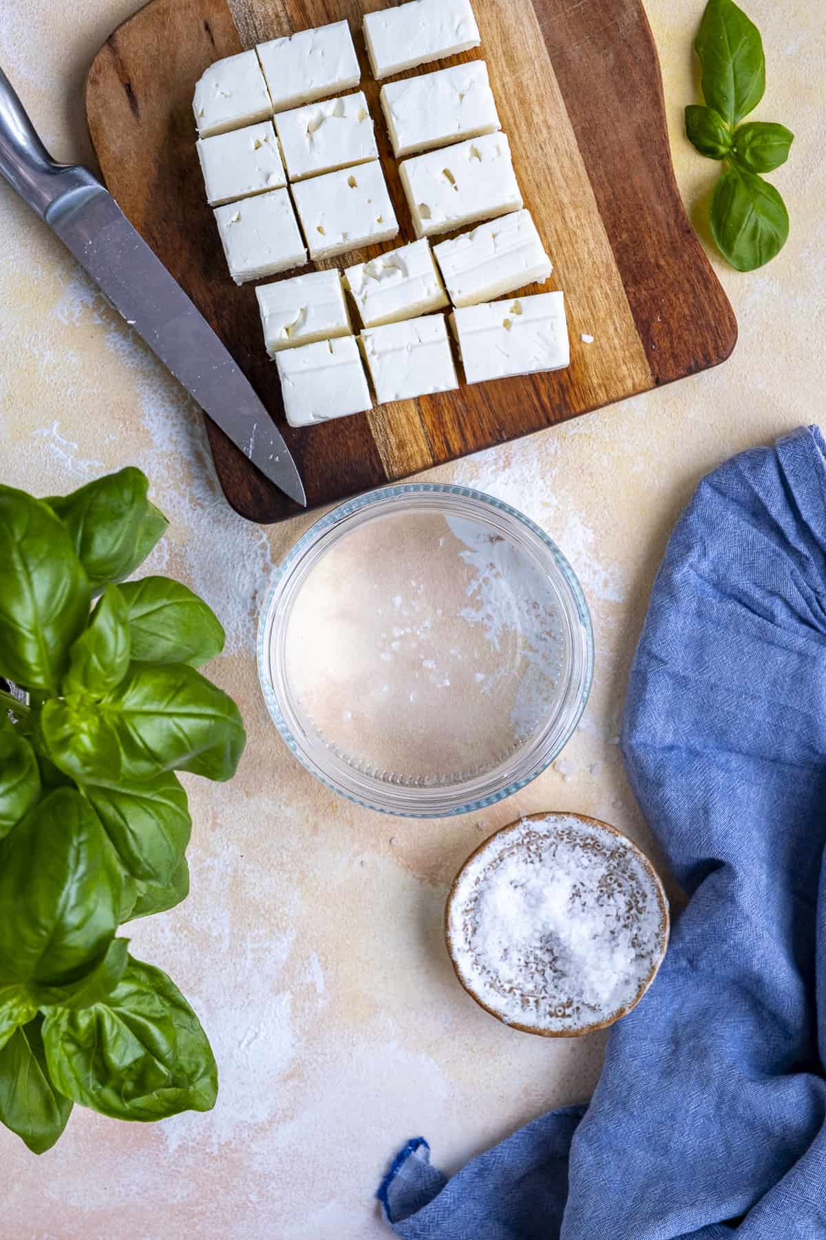Water in a jar, kosher salt in a mini bowl, feta cheese cubes on a cutting board and fresh basil photographed on a light background.