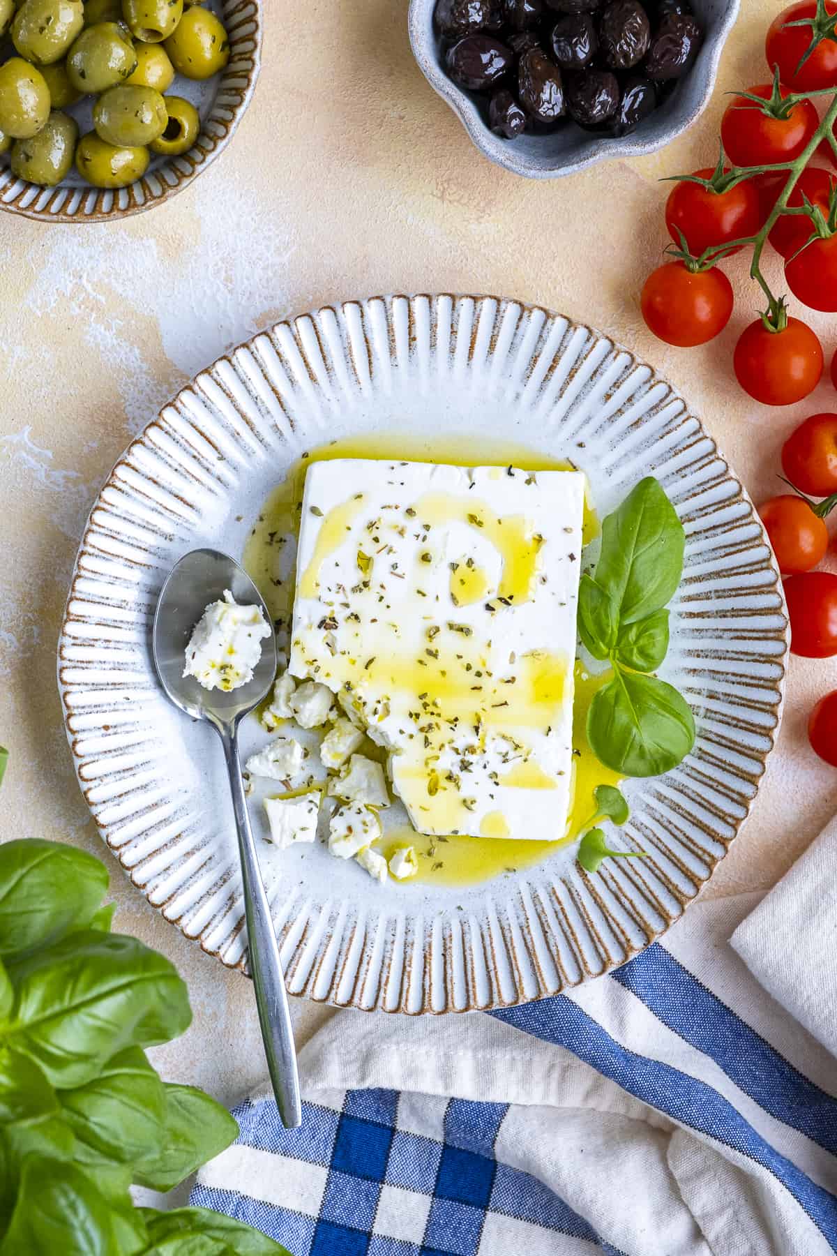 A block of feta cheese seasoned with oregano and drizzled with olive oil on a white plate. Olives, tomatoes and fresh basil on the side.