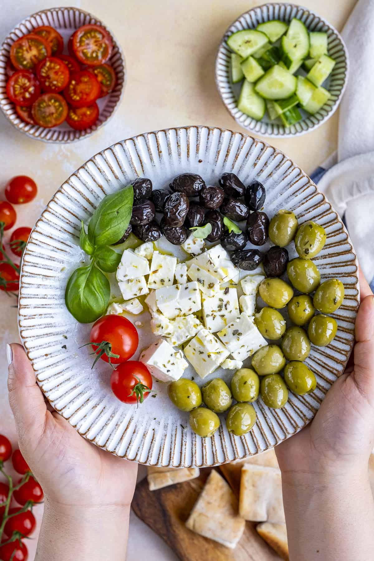 Hands holding a plate full of black olives, green olives, feta cheese and tomatoes.