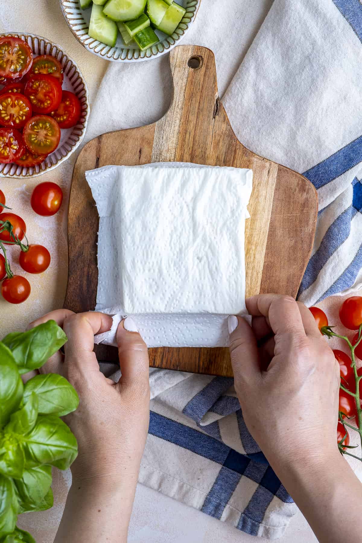 Hands wrapping a block of feta cheese with dampened paper towel. Sliced tomatoes, cucumber and fresh basil on the side.