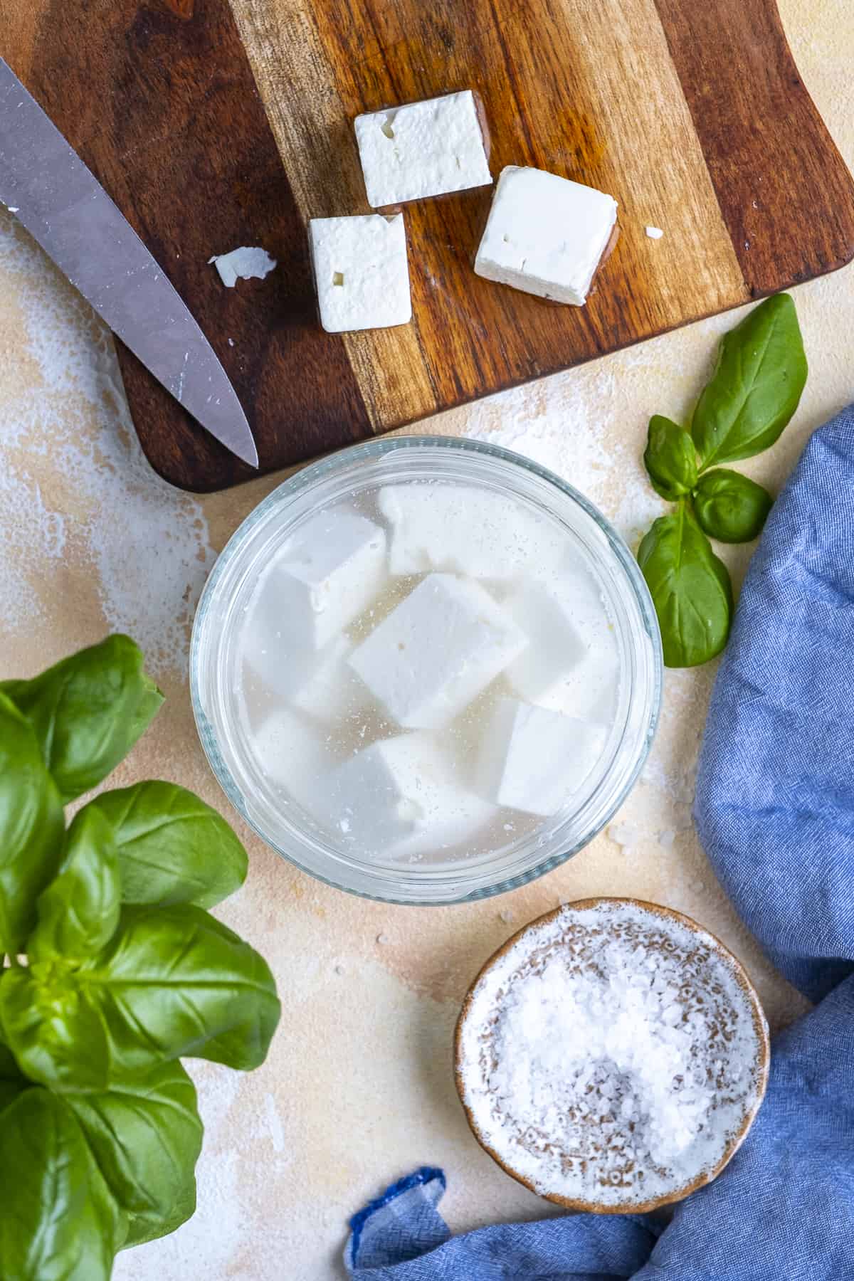 Feta cheese cubes in brine, kosher salt, more cheese on a cutting board and fresh basil on the side.