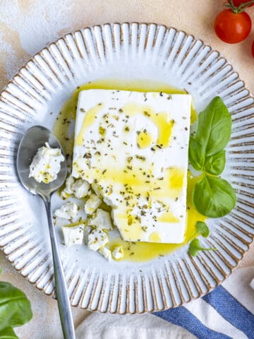A block of feta cheese, one corner of it crumbled with a spoon on a white plate.