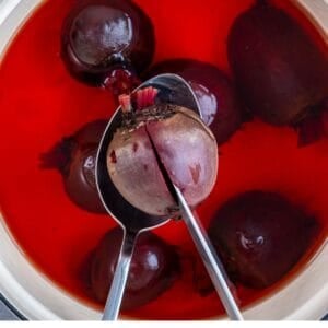 Whole beets boiling in a pot with deep red water, one beet lifted with a spoon and pierced with a knife to check tenderness.