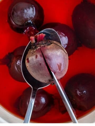 Whole beets boiling in a pot with deep red water, one beet lifted with a spoon and pierced with a knife to check tenderness.