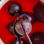 Whole beets boiling in a pot with deep red water, one beet lifted with a spoon and pierced with a knife to check tenderness.