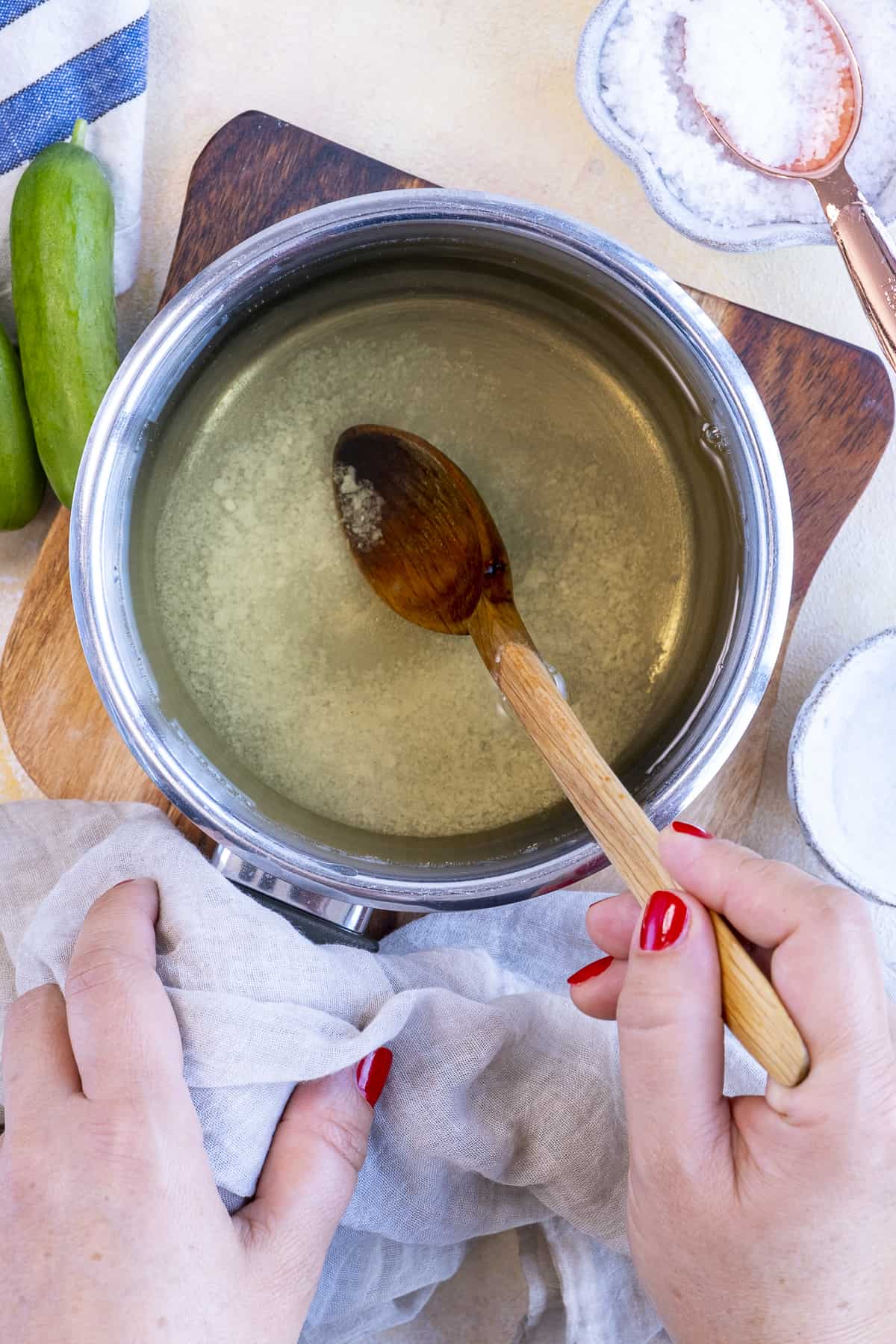 Hands mixing pickle brine with a wooden spoon in a saucepan.