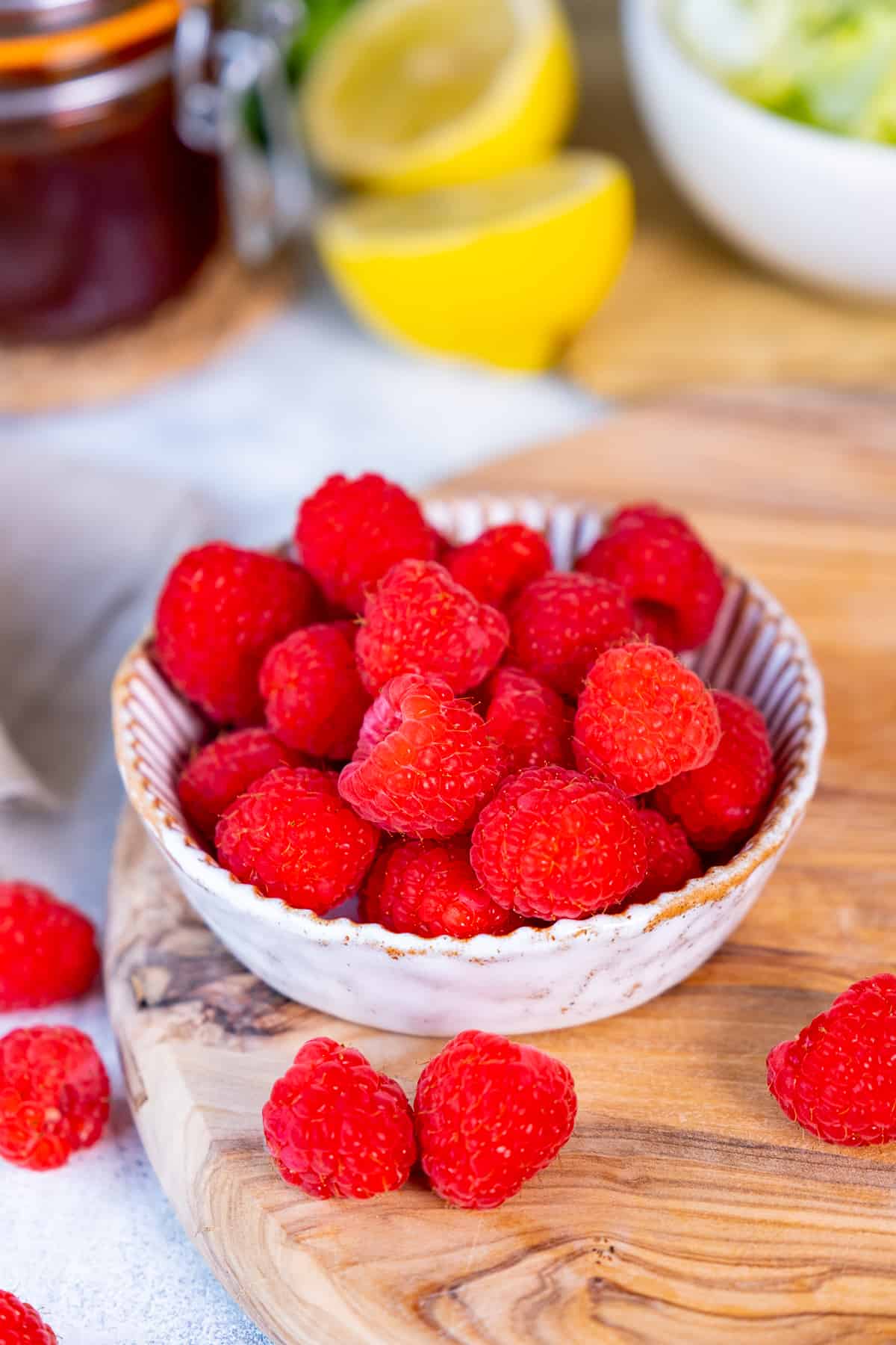 Fresh raspberries in a white bowl on a wooden board, lemons, a jam jar and a salad bowl behind them.