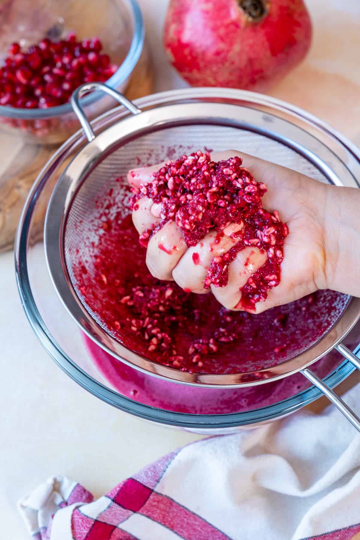 A hand squeezing pomegranate arils over a fine mesh sieve.