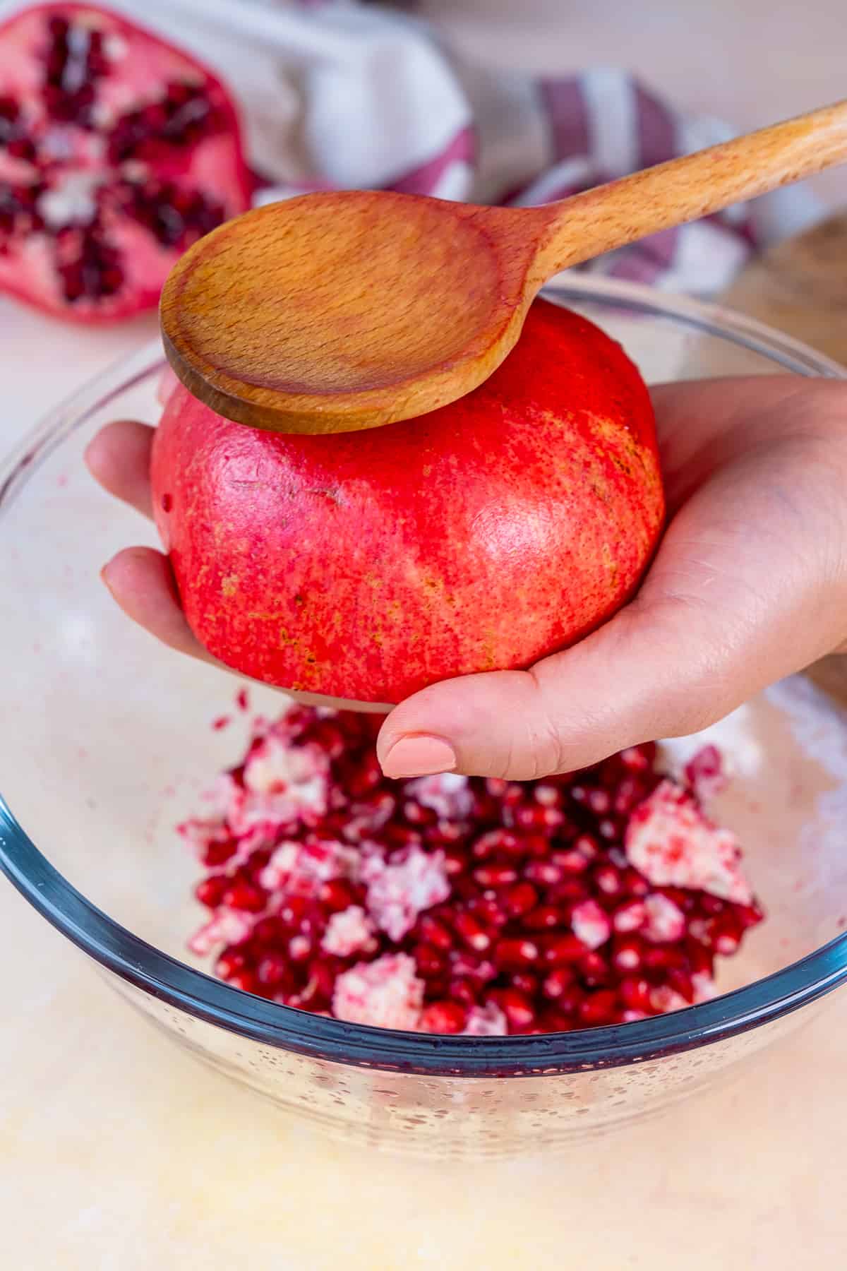 Hands smacking a half pomegranate with a wooden spoon over a mixing bowl.