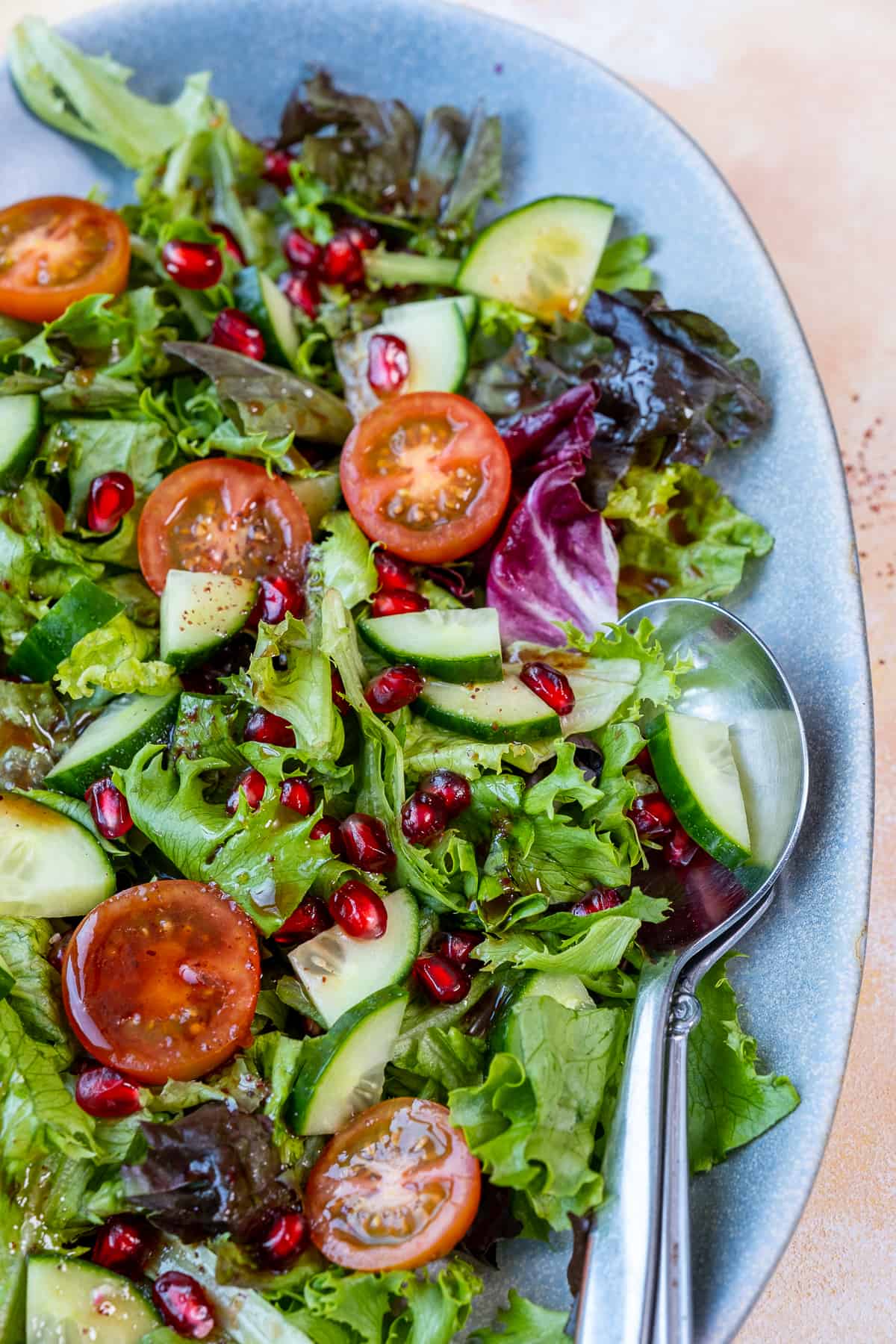 A green salad with lettuce, tomatoes, cucumbers and pomegranate arils in an oval dish and two spoons in it.