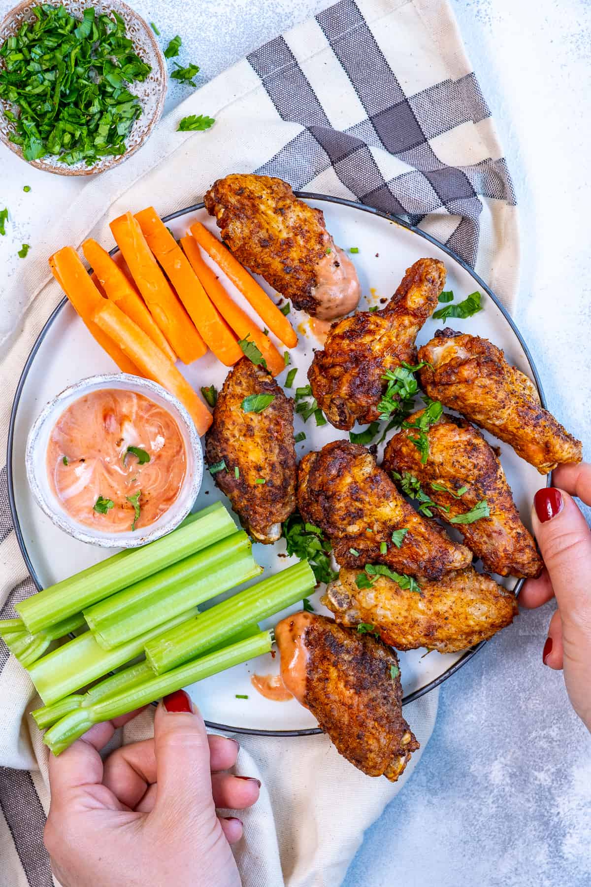 Hands holding a plate loaded with chicken wings, celery sticks, carrot sticks and a dipping sauce.