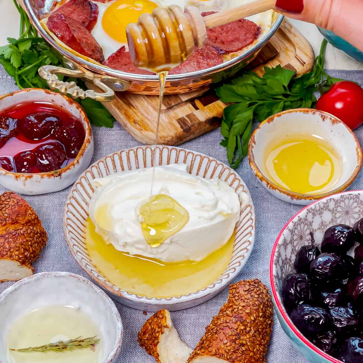 Honey is being drizzled over kaymak in a small bowl, other breakfast foods around it.