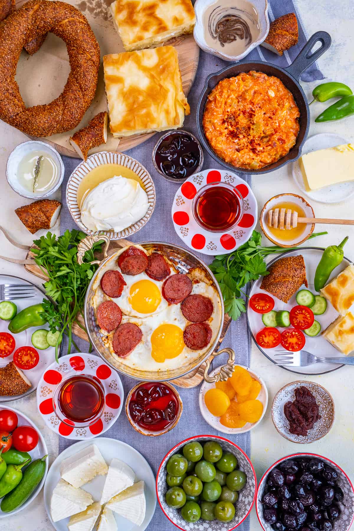 Flat-lay of Turkish breakfast with egg dishes in copper pan, pastries like borek and simit, jams, olives, cheese, vegetables and Turkish tea in traditional tulip glasses.