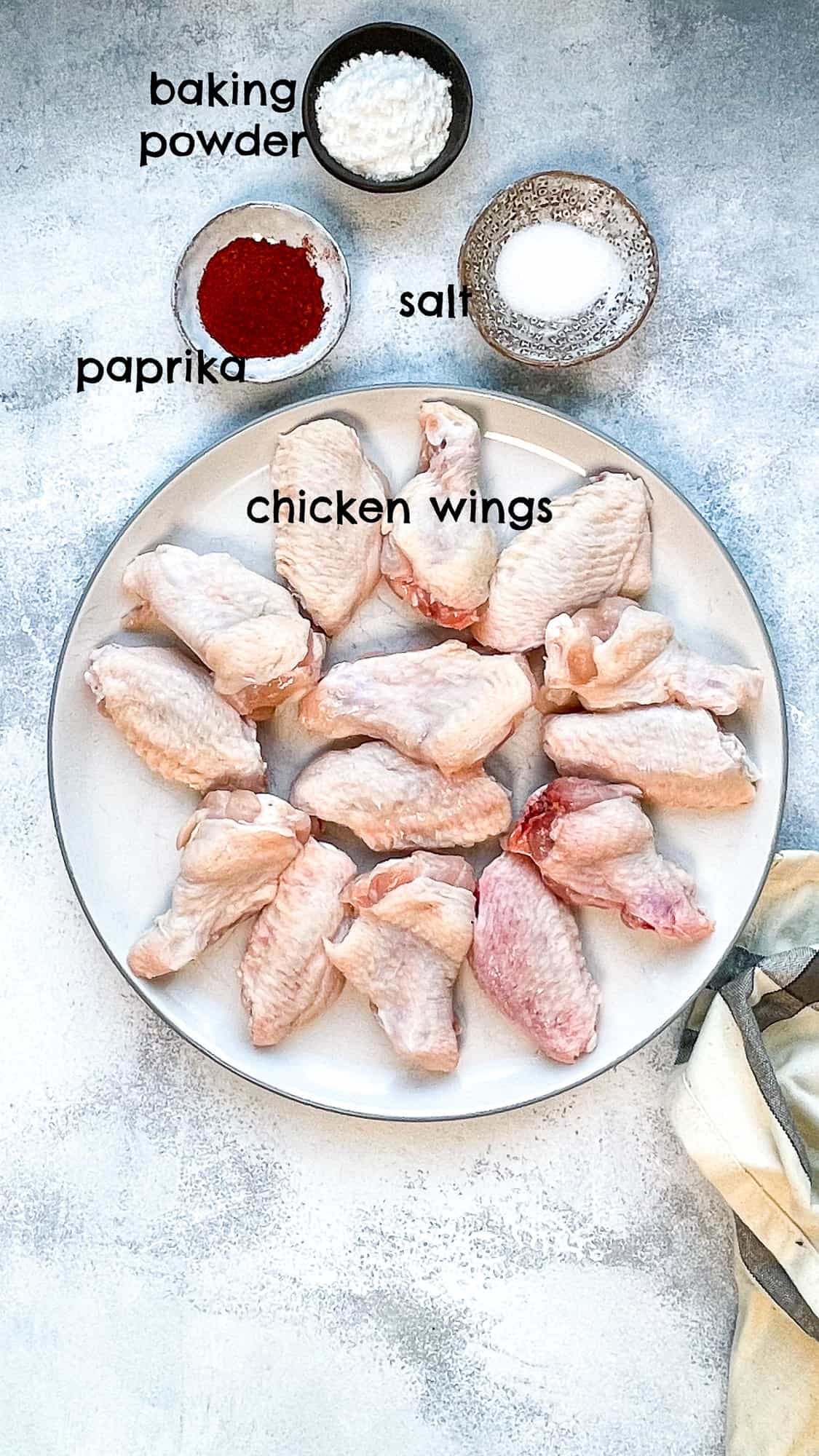 Raw chicken wings on a plate, paprika, salt and baking powder in small bowls around it.