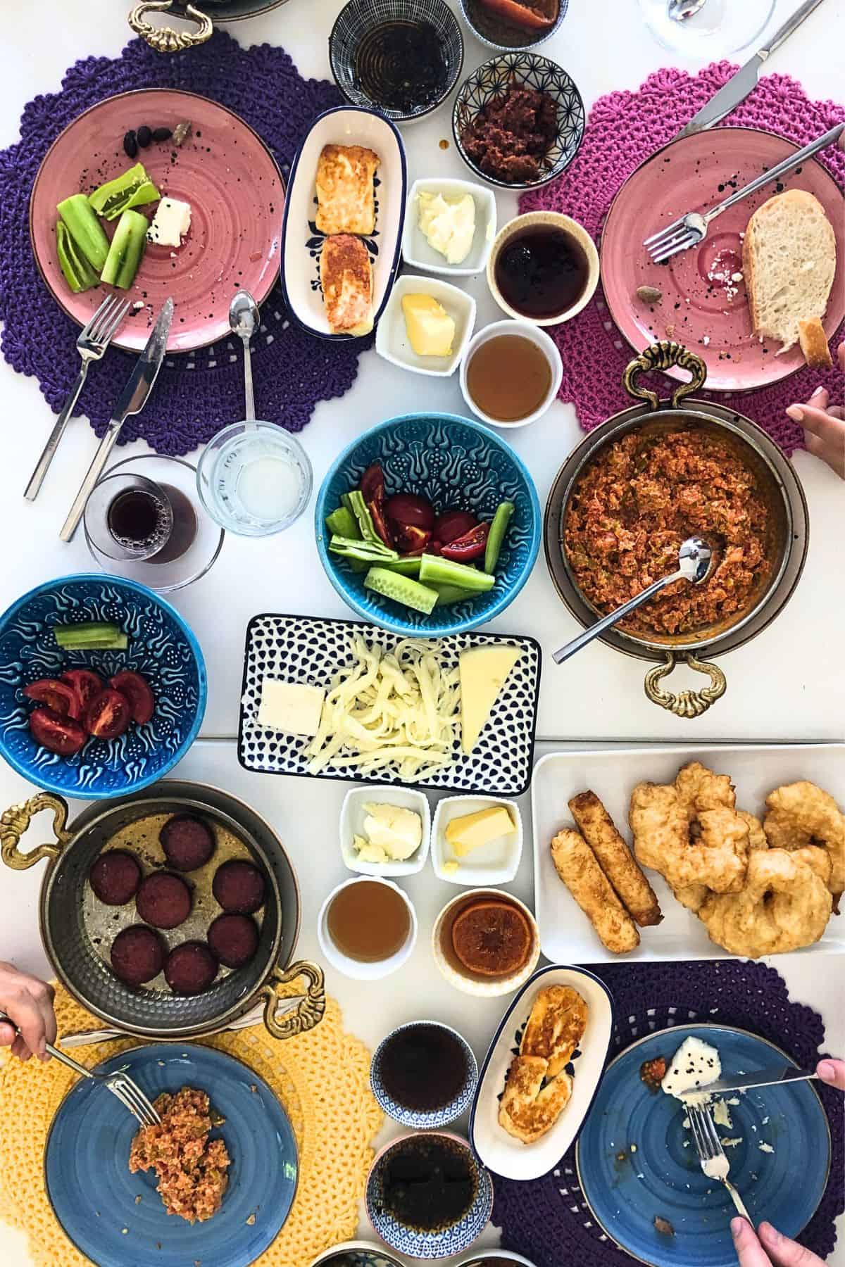 Traditional Turkish breakfast table with menemen, fried eggs with sucuk, cheeses, olives, fresh vegetables, fried dough, bread, and Turkish tea served in small glasses.
