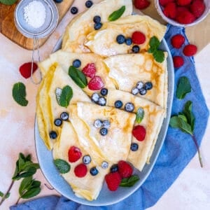 Crepes served with fresh berries in an oval plate. Powdered sugar and mint leaves accompany.