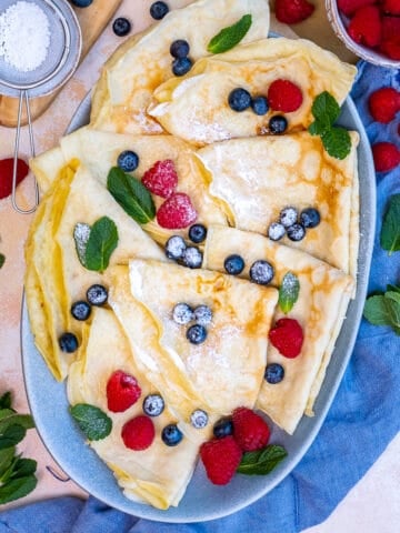 Crepes served with fresh berries in an oval plate. Powdered sugar and mint leaves accompany.