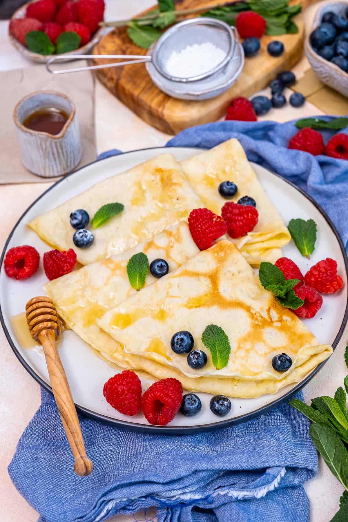 Crepes served with fresh berries, mint leaves and maple syrup on a white plate.