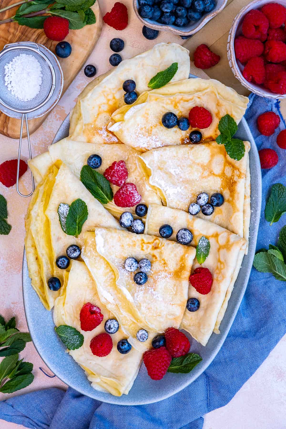 Crepes served with a little powdered sugar, fresh berries and mint leaves on an oval plate.