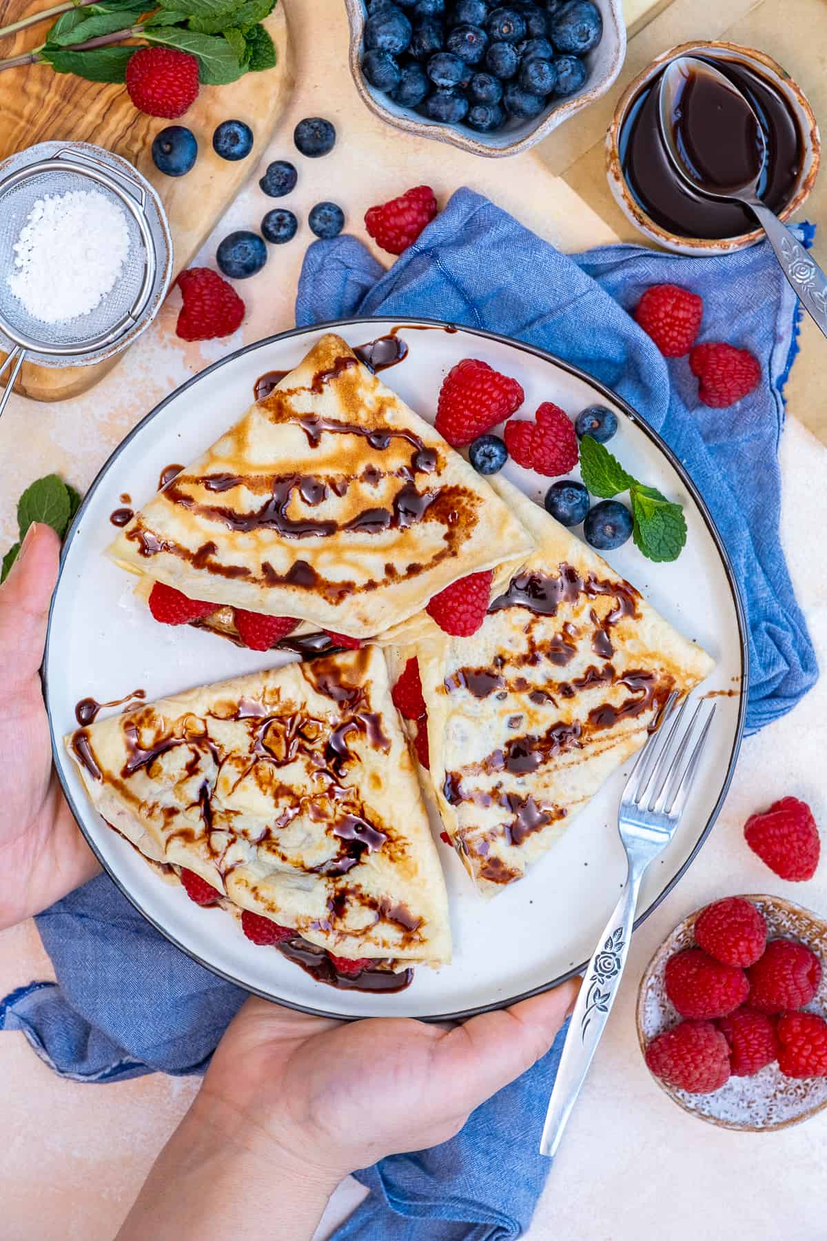 Woman holding a plate with crepes filled and topped with chocolate and berries.