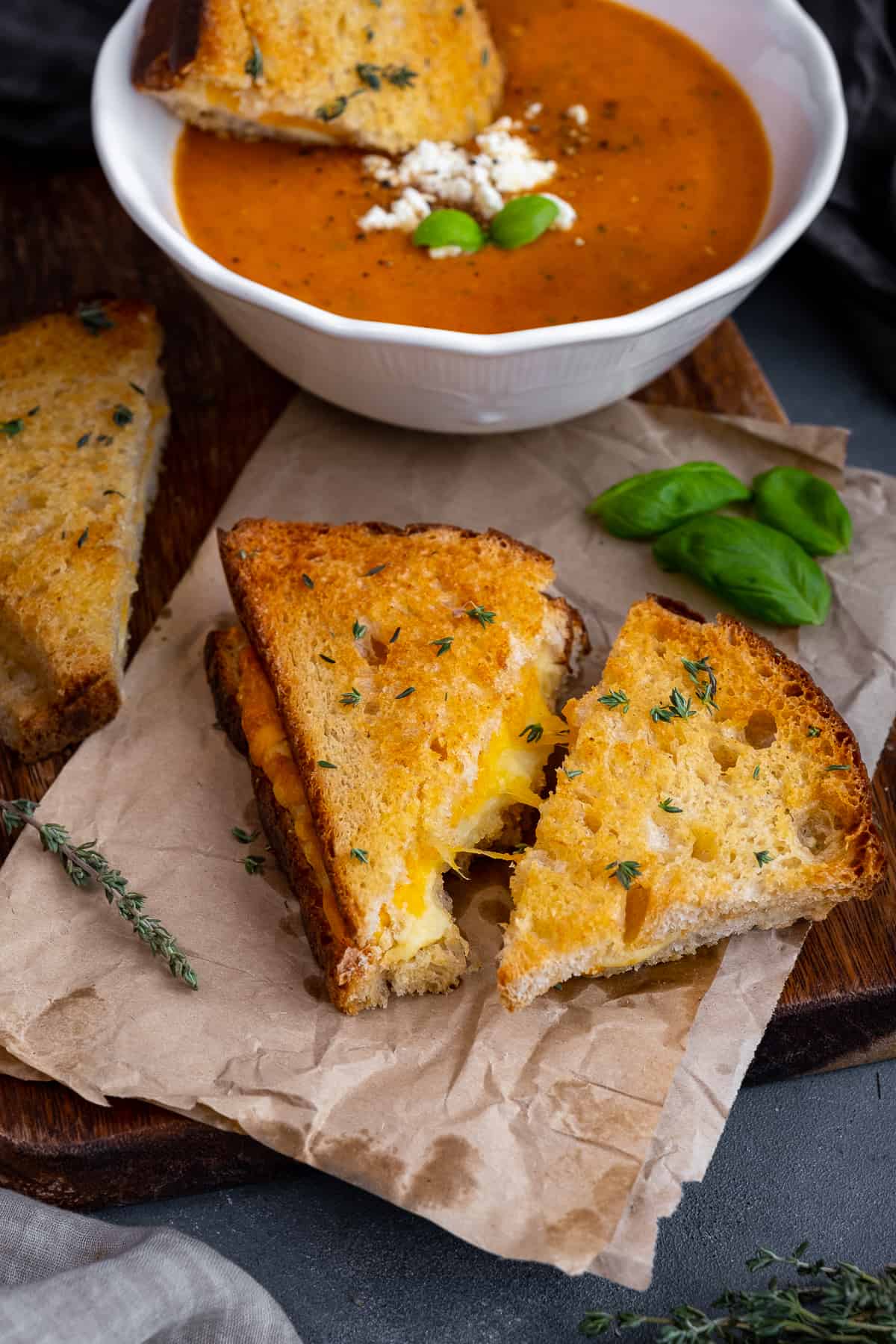 Grilled cheese sandwiches garnished with thyme and a bowl of tomato soup behind them.