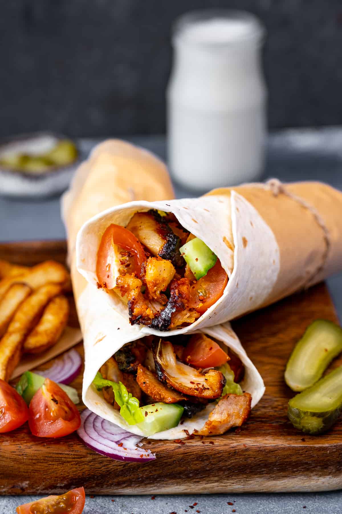 Chicken doner wraps accompanied by French fries, pickles, sliced tomatoes and onions on a wooden board and a glass of ayran behind these.