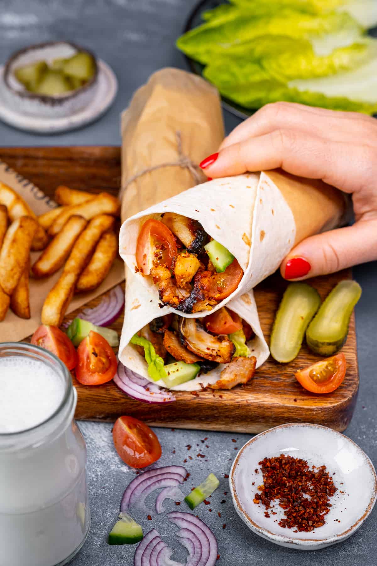 A hand taking a chicken doner wrap from a wooden board loaded with chips, pickles, tomatoes and onions.