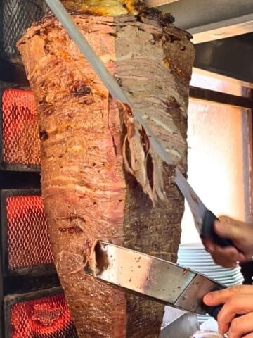 Close-up of beef doner kebab being sliced from a vertical rotisserie, showing layers of meat and fat as it cooks.