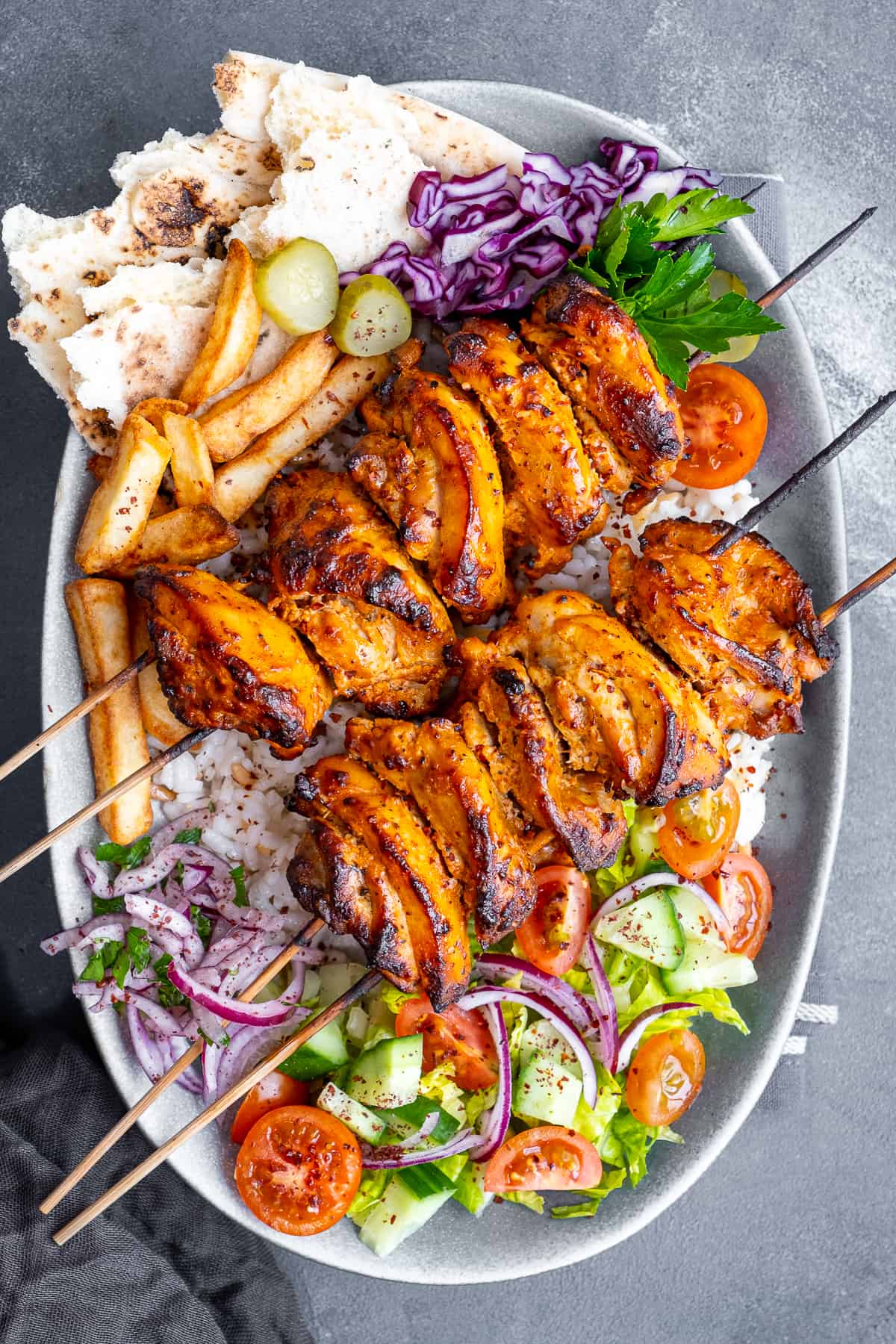 Chicken doner kebab skewers accompanied by rice, salad, pickles and French fries on the same oval plate.