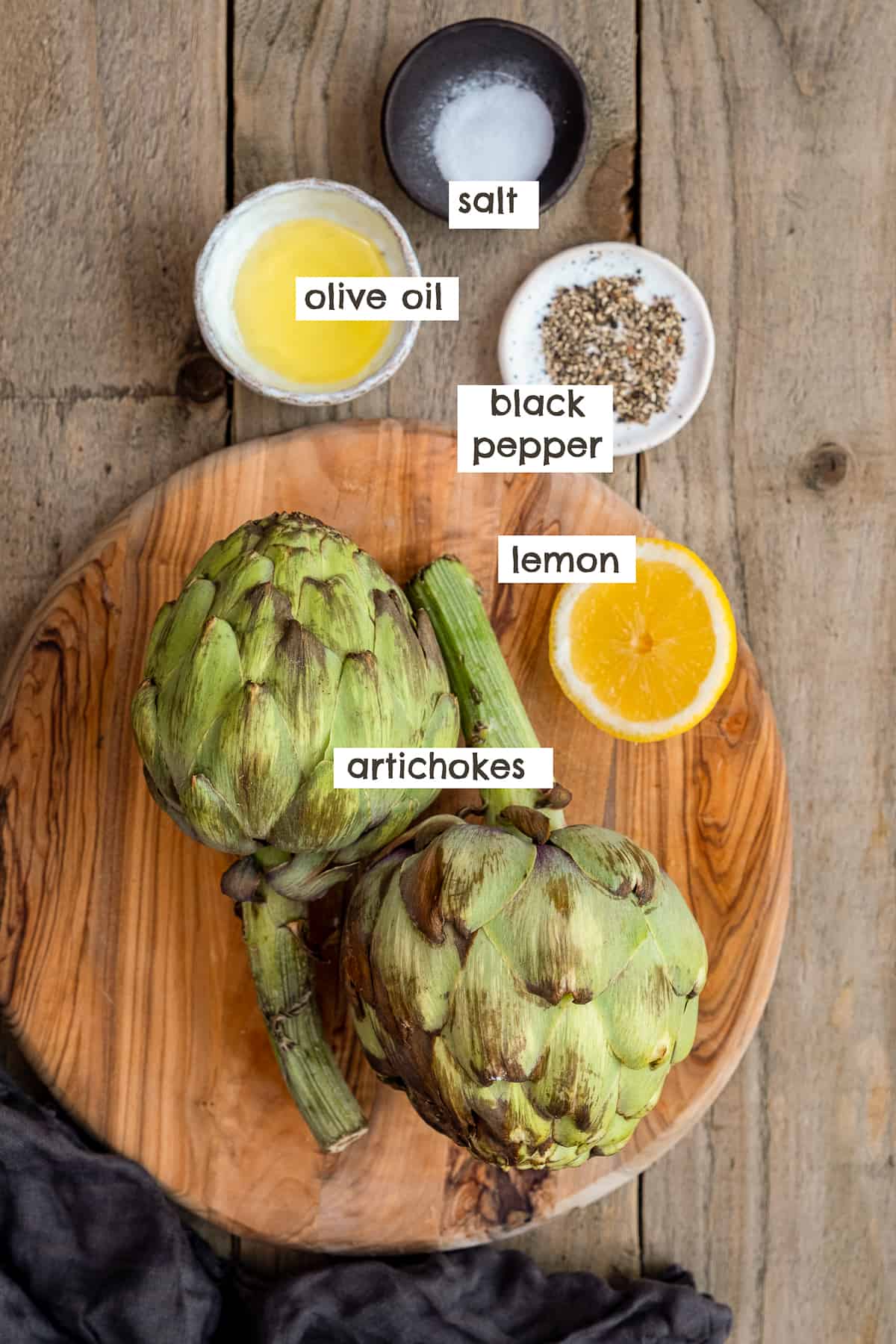 Artichokes, half lemon, black pepper, salt and olive oil on a wooden board.