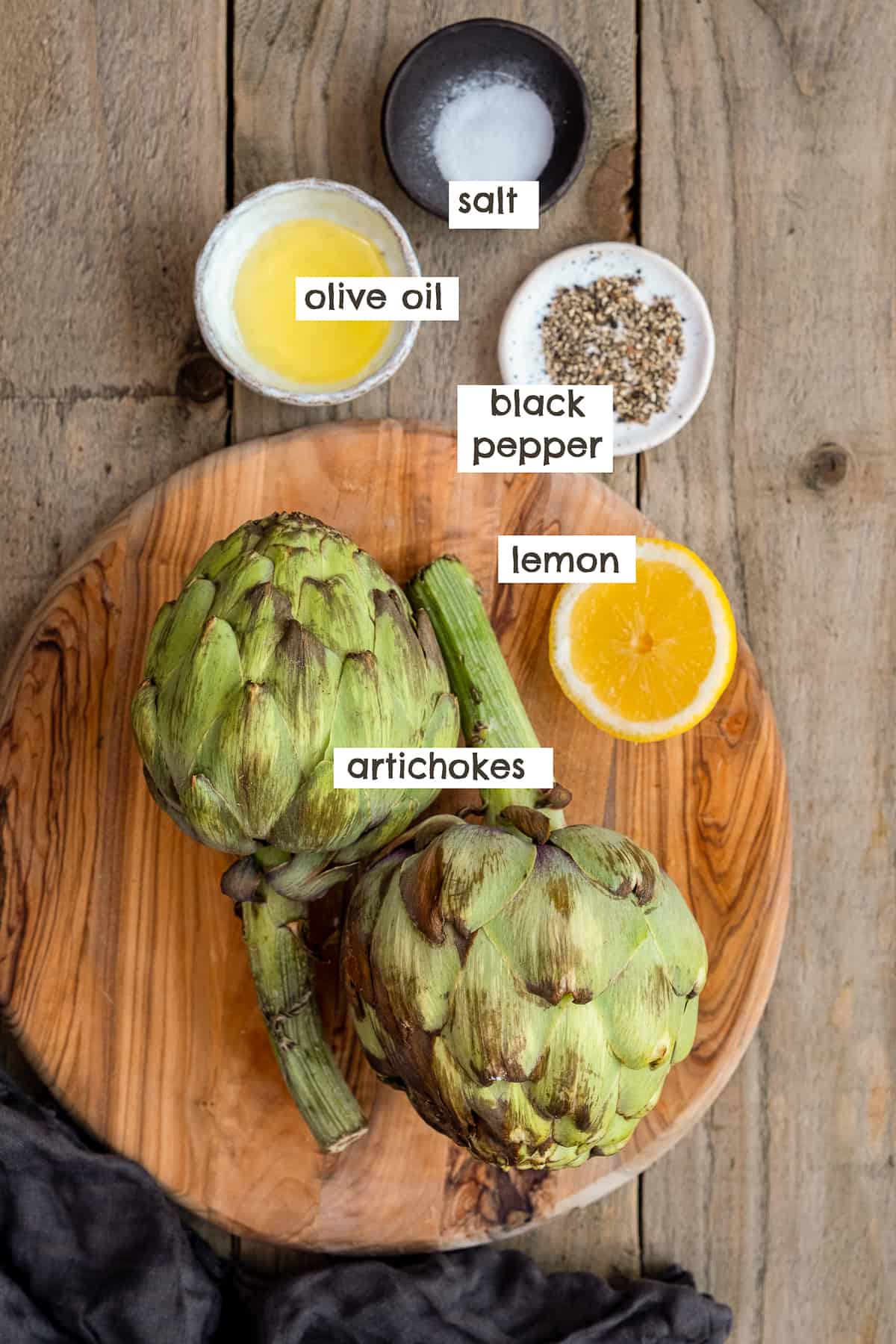 Artichokes, half lemon, black pepper, salt and olive oil on a wooden board.