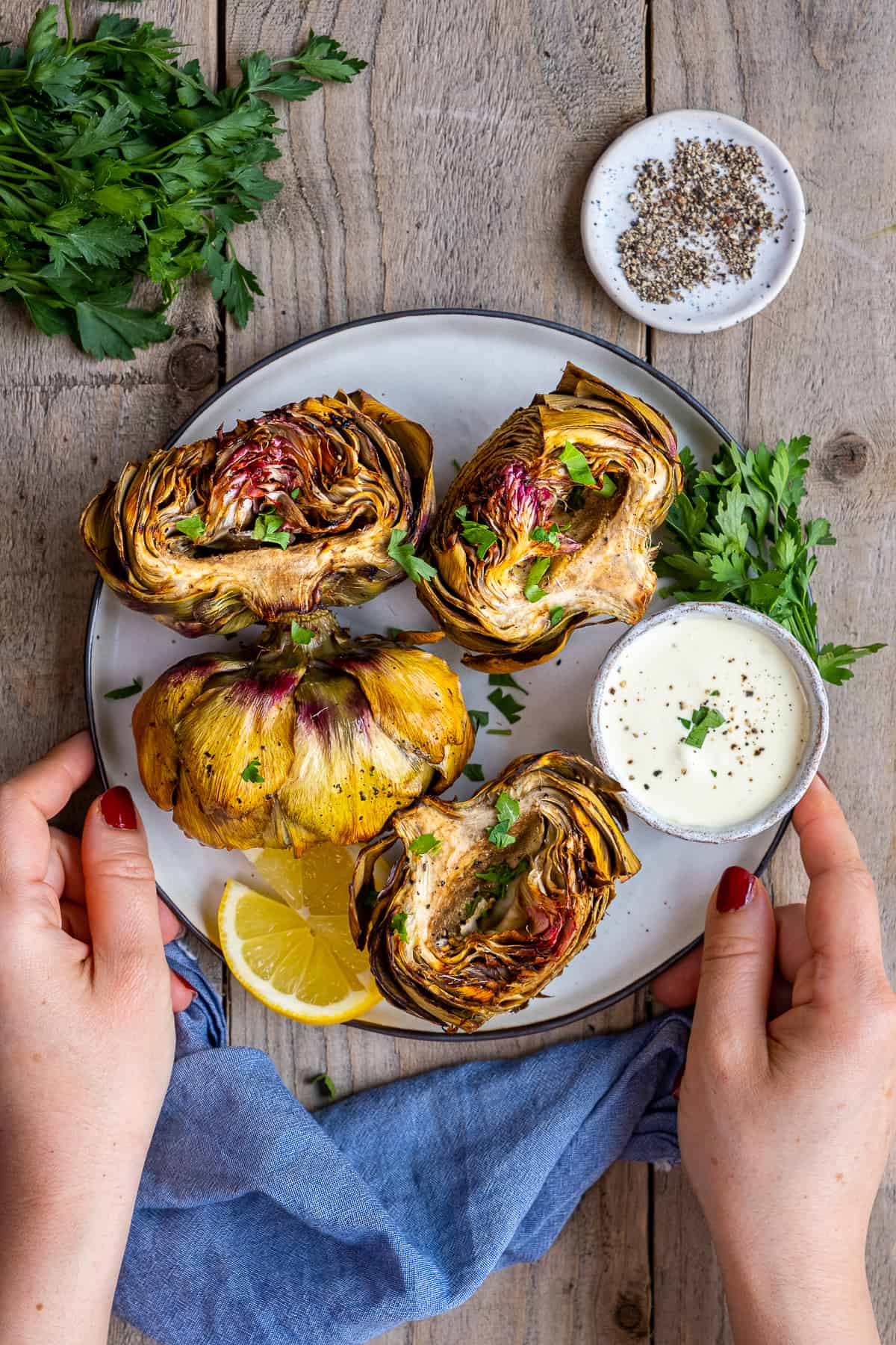 Woman hands holding a plate filled with air fried artichokes, lemon slices, parsley and a dipping sauce in a mini bowl.