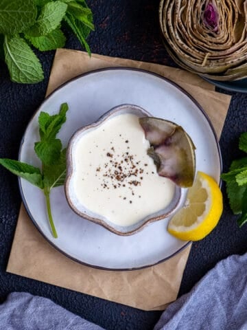 Dipping sauce in a small ceramic bowl, topped with ground black pepper and artichoke leaves, a slice of lemon and fresh mint on the side.