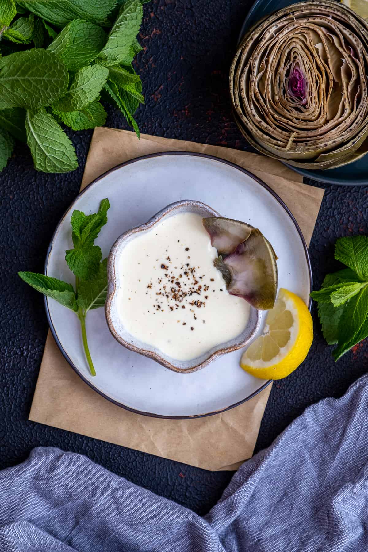 A bowl of artichoke dipping sauce garnished with black pepper and artichoke leaves on the side photographed from top view.