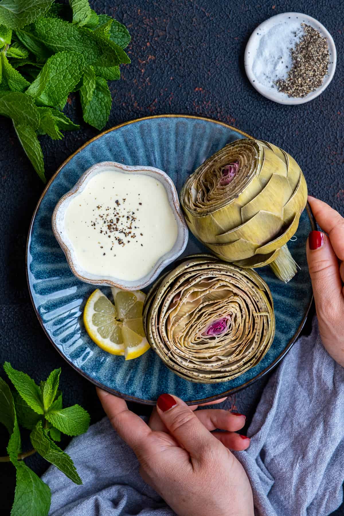 Woman serving steamed artichokes paired with a bowl of dipping sauce on a dark blue plate.