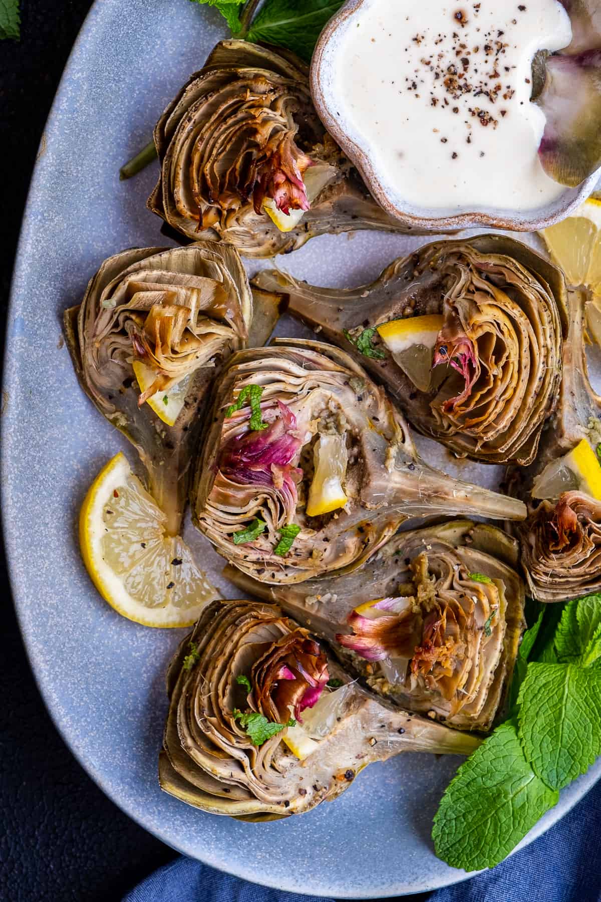 Oven roasted artichokes served on an oval plate with a bowl of dipping sauce on the side.