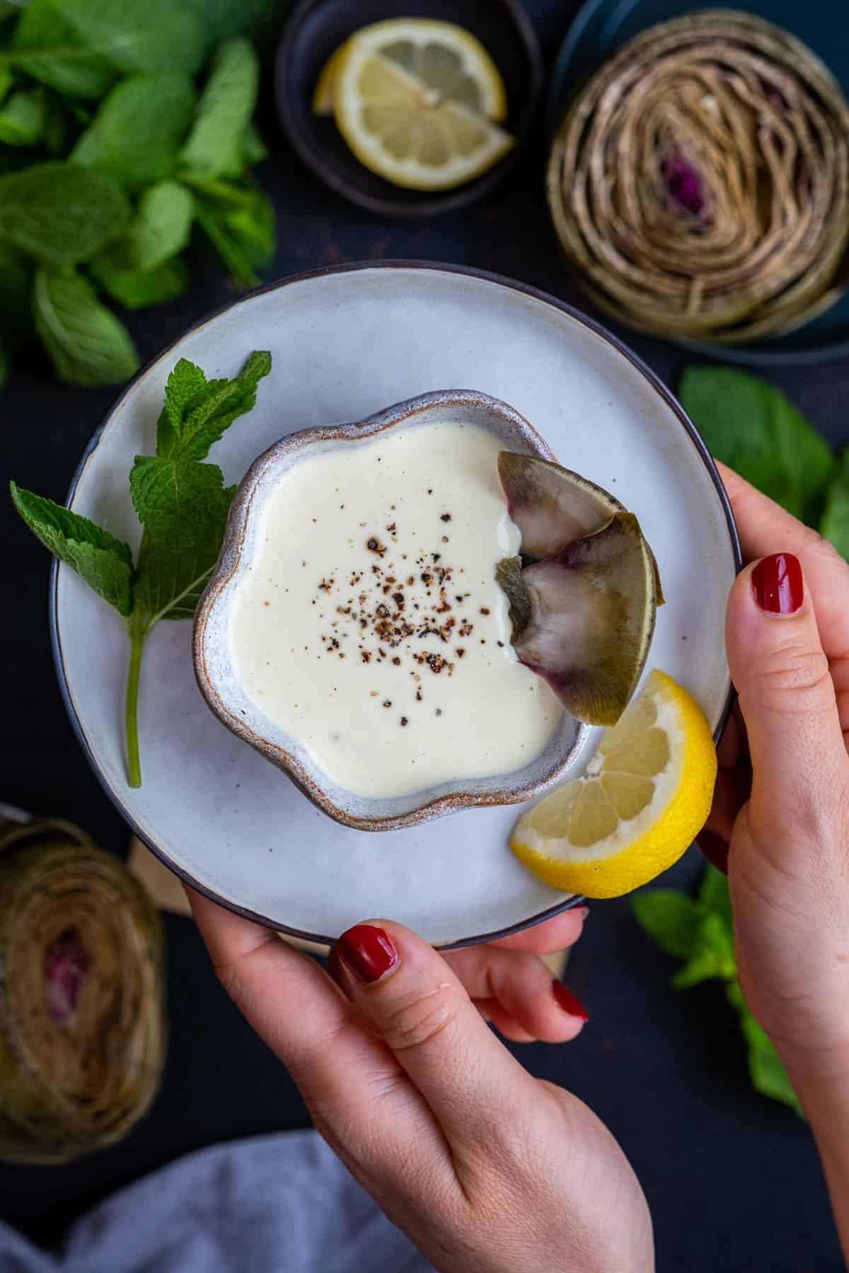 Woman hands serving a bowl of artichoke dipping sauce placed on a plate.