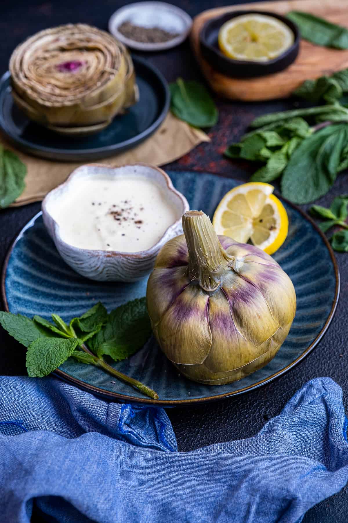 A steamed artichoke served with a bowl of yogurt and mayonnaise dipping sauce on a dark blue plate.