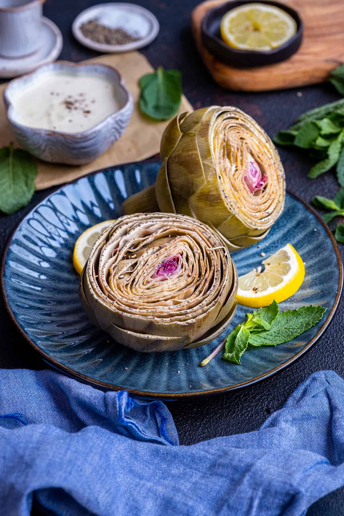 Steamed artichokes garnished with lemon slices and fresh mint leaves on a dark blue plate photographed from front view.