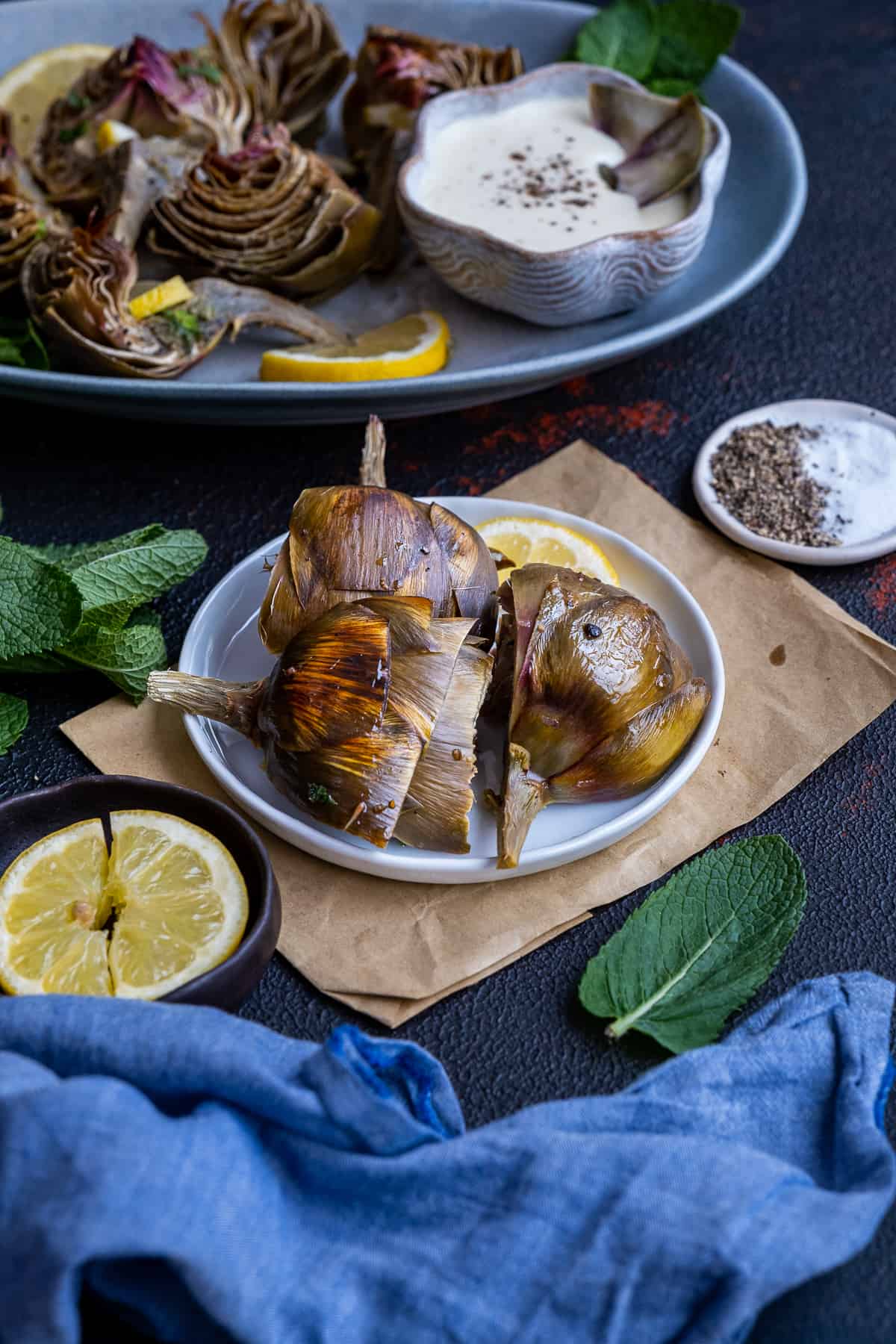 Roasted artichokes served on a white plate, accompanied by a bowl of dipping sauce, mint leaves and lemon slices photographed from front view.