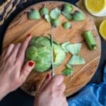 Woman hands cutting off the top of an artichoke on a wooden board.